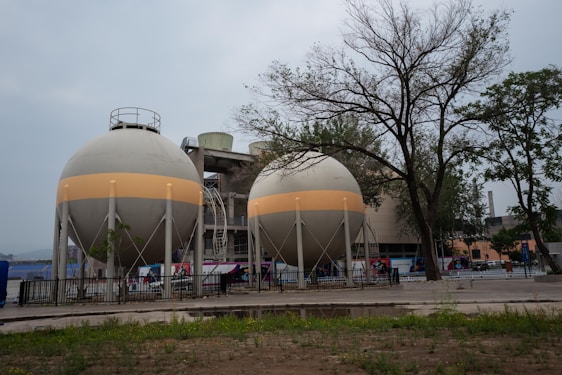 Two large, spherical industrial tanks are situated in an outdoor area, surrounded by a fence. These tanks are supported by multiple legs and feature a horizontal beige band around their circumference. In the background, there are trees with sparse foliage and a larger industrial building. The foreground consists of patches of grass and bare ground.