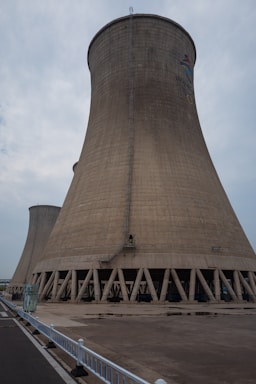 A large cooling tower stands prominently against a cloudy sky. The structure is made of concrete and features a flared base, characterized by a series of vertical support beams around its bottom perimeter. A small ladder runs vertically along the side, indicating human access for maintenance purposes. The ground in front is paved, with a white fence lining the edge of a road on the left side.