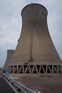 A large cooling tower stands prominently against a cloudy sky. The structure is made of concrete and features a flared base, characterized by a series of vertical support beams around its bottom perimeter. A small ladder runs vertically along the side, indicating human access for maintenance purposes. The ground in front is paved, with a white fence lining the edge of a road on the left side.