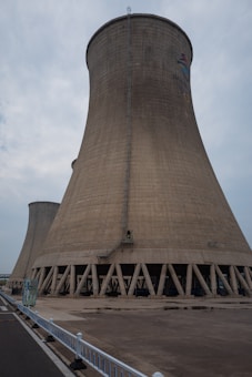 A large cooling tower stands prominently against a cloudy sky. The structure is made of concrete and features a flared base, characterized by a series of vertical support beams around its bottom perimeter. A small ladder runs vertically along the side, indicating human access for maintenance purposes. The ground in front is paved, with a white fence lining the edge of a road on the left side.