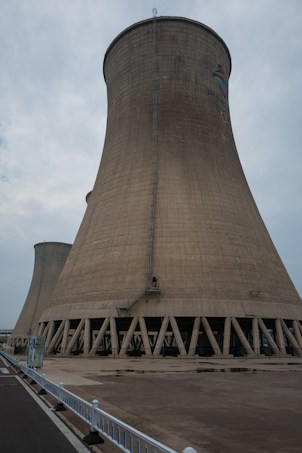 A large cooling tower stands prominently against a cloudy sky. The structure is made of concrete and features a flared base, characterized by a series of vertical support beams around its bottom perimeter. A small ladder runs vertically along the side, indicating human access for maintenance purposes. The ground in front is paved, with a white fence lining the edge of a road on the left side.