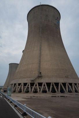 A large cooling tower stands prominently against a cloudy sky. The structure is made of concrete and features a flared base, characterized by a series of vertical support beams around its bottom perimeter. A small ladder runs vertically along the side, indicating human access for maintenance purposes. The ground in front is paved, with a white fence lining the edge of a road on the left side.