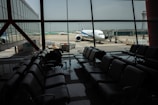 The image depicts an airport terminal with empty seating areas facing large windows through which a stationary airplane is visible on the tarmac. The airport environment appears quiet and subdued, with soft lighting casting reflections on the shiny floor. A luggage conveyor can be seen outside near the airplane, and there is a control tower visible in the background.