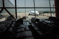 The image depicts an airport terminal with empty seating areas facing large windows through which a stationary airplane is visible on the tarmac. The airport environment appears quiet and subdued, with soft lighting casting reflections on the shiny floor. A luggage conveyor can be seen outside near the airplane, and there is a control tower visible in the background.