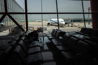 The image depicts an airport terminal with empty seating areas facing large windows through which a stationary airplane is visible on the tarmac. The airport environment appears quiet and subdued, with soft lighting casting reflections on the shiny floor. A luggage conveyor can be seen outside near the airplane, and there is a control tower visible in the background.