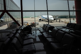 The image depicts an airport terminal with empty seating areas facing large windows through which a stationary airplane is visible on the tarmac. The airport environment appears quiet and subdued, with soft lighting casting reflections on the shiny floor. A luggage conveyor can be seen outside near the airplane, and there is a control tower visible in the background.