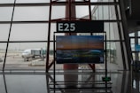 A jet bridge connects to an airplane at a gate marked E25 in an airport terminal. The terminal interior features large windows that provide a view of the aircraft and outdoor tarmac. A digital sign displays flight information, including a departure to Taipei at 13:55.