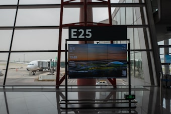 A jet bridge connects to an airplane at a gate marked E25 in an airport terminal. The terminal interior features large windows that provide a view of the aircraft and outdoor tarmac. A digital sign displays flight information, including a departure to Taipei at 13:55.