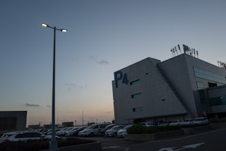 A parking lot during dusk with numerous parked cars adjacent to a large, modern building labeled P4. The sky is clear, with a blue gradient transitioning to warm hues near the horizon. Streetlights are illuminated, adding a soft glow to the scene.