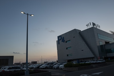 A parking lot during dusk with numerous parked cars adjacent to a large, modern building labeled P4. The sky is clear, with a blue gradient transitioning to warm hues near the horizon. Streetlights are illuminated, adding a soft glow to the scene.