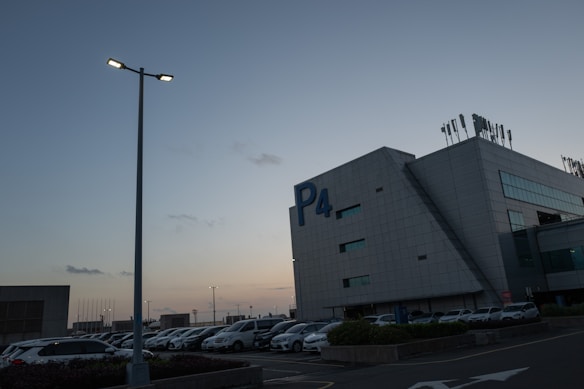 A parking lot during dusk with numerous parked cars adjacent to a large, modern building labeled P4. The sky is clear, with a blue gradient transitioning to warm hues near the horizon. Streetlights are illuminated, adding a soft glow to the scene.