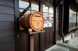 A wooden barrel-shaped mailbox with a slot and emblem on the side is placed on a stand outside a traditional wooden building with large windows. The building exterior is made of dark wood, and the window panes reflect light. The scene appears to be part of a quiet, traditional area, with minimal visible activity.