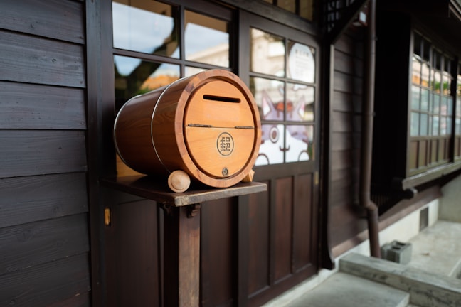 A wooden barrel-shaped mailbox with a slot and emblem on the side is placed on a stand outside a traditional wooden building with large windows. The building exterior is made of dark wood, and the window panes reflect light. The scene appears to be part of a quiet, traditional area, with minimal visible activity.
