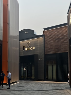 Modern architectural setting featuring a building with the sign 'Puerto 99'. The structure has a mix of dark panels and wood finishes. Two people are standing on the left, engaged in conversation or looking at a phone. The ground is paved, suggesting a commercial or urban area.