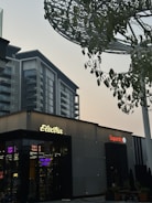 A modern urban scene featuring a commercial building with signs for 'Electra' and 'Superdry'. The architecture is contemporary, with a visible high-rise building in the backdrop. There are trees with green leaves and a large metallic structure resembling a canopy or art installation above.