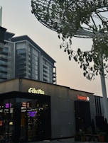 A modern urban scene featuring a commercial building with signs for 'Electra' and 'Superdry'. The architecture is contemporary, with a visible high-rise building in the backdrop. There are trees with green leaves and a large metallic structure resembling a canopy or art installation above.