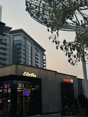 A modern urban scene featuring a commercial building with signs for 'Electra' and 'Superdry'. The architecture is contemporary, with a visible high-rise building in the backdrop. There are trees with green leaves and a large metallic structure resembling a canopy or art installation above.
