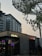 A modern urban scene featuring a commercial building with signs for 'Electra' and 'Superdry'. The architecture is contemporary, with a visible high-rise building in the backdrop. There are trees with green leaves and a large metallic structure resembling a canopy or art installation above.