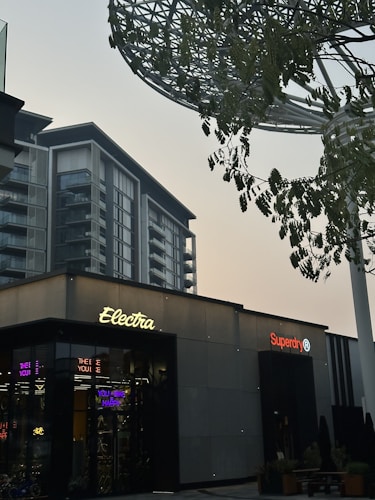 A modern urban scene featuring a commercial building with signs for 'Electra' and 'Superdry'. The architecture is contemporary, with a visible high-rise building in the backdrop. There are trees with green leaves and a large metallic structure resembling a canopy or art installation above.