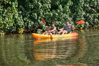 Two people are kayaking on a calm body of water surrounded by dense green foliage. They are both wearing sunglasses and hats, and appear to be enjoying their outdoor adventure. The kayak is orange and yellow, and the water reflects the lush greenery and the kayak's colors.
