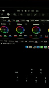 The image shows a computer screen displaying a video editing software interface, specifically DaVinci Resolve 18.5. Various color adjustment wheels for shadows, midtones, and highlights are visible, along with multiple control panels and icons at the top. A keyboard is partially visible at the bottom.