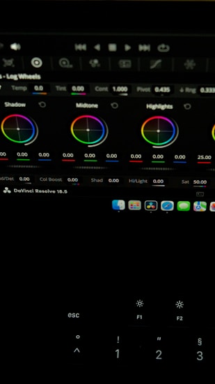 The image shows a computer screen displaying a video editing software interface, specifically DaVinci Resolve 18.5. Various color adjustment wheels for shadows, midtones, and highlights are visible, along with multiple control panels and icons at the top. A keyboard is partially visible at the bottom.