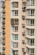 Close-up view of two modern apartment buildings, featuring multiple balconies and large windows. The buildings exhibit an urban architectural style, with one being light peach and the other in light beige. Metal structures on the balconies indicate the presence of air conditioning units.