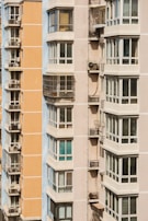 Close-up view of two modern apartment buildings, featuring multiple balconies and large windows. The buildings exhibit an urban architectural style, with one being light peach and the other in light beige. Metal structures on the balconies indicate the presence of air conditioning units.