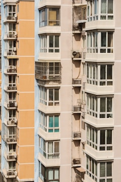 Close-up view of two modern apartment buildings, featuring multiple balconies and large windows. The buildings exhibit an urban architectural style, with one being light peach and the other in light beige. Metal structures on the balconies indicate the presence of air conditioning units.