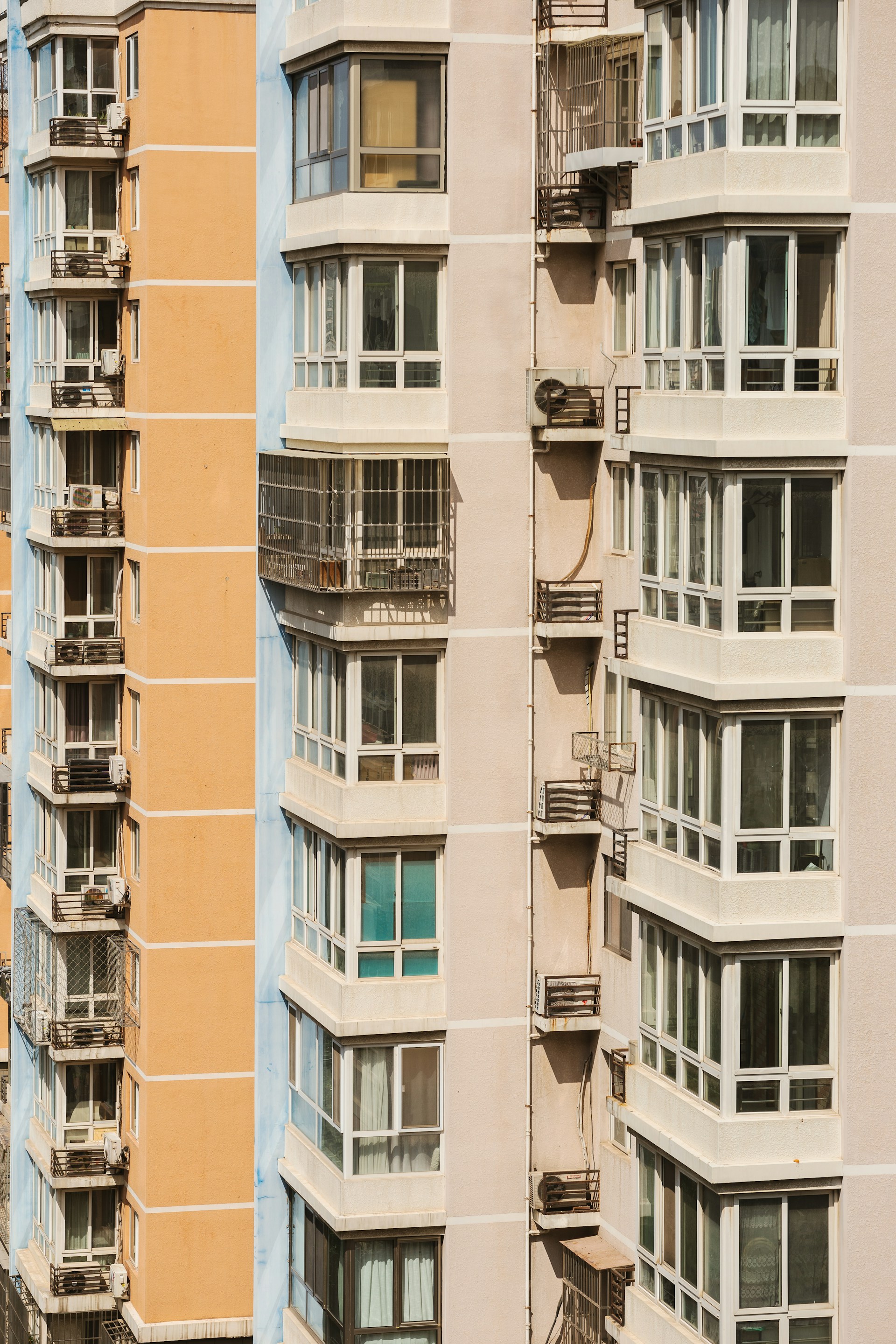 Close-up view of two modern apartment buildings, featuring multiple balconies and large windows. The buildings exhibit an urban architectural style, with one being light peach and the other in light beige. Metal structures on the balconies indicate the presence of air conditioning units.