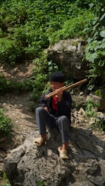 A young person sitting on a rock surrounded by lush green foliage, playing a wooden flute. The person is dressed in casual clothing, including a hat and slippers, exuding a serene and natural ambiance.