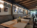 A cozy restaurant interior with a row of long benches and wooden tables set with placemats. The walls are adorned with various framed pictures and round lights. The ceiling features exposed wooden beams, and large glass doors let in natural light.