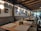 A cozy restaurant interior with a row of long benches and wooden tables set with placemats. The walls are adorned with various framed pictures and round lights. The ceiling features exposed wooden beams, and large glass doors let in natural light.