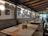 A cozy restaurant interior with a row of long benches and wooden tables set with placemats. The walls are adorned with various framed pictures and round lights. The ceiling features exposed wooden beams, and large glass doors let in natural light.
