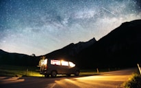 A camper van is parked on a roadside under a clear night sky filled with stars and a visible Milky Way. The van's interior lights are on, casting a warm glow, and a bicycle is attached to the back. Mountains create a silhouetted backdrop, adding to the serene and adventurous setting.