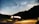 A camper van is parked on a roadside under a clear night sky filled with stars and a visible Milky Way. The van's interior lights are on, casting a warm glow, and a bicycle is attached to the back. Mountains create a silhouetted backdrop, adding to the serene and adventurous setting.