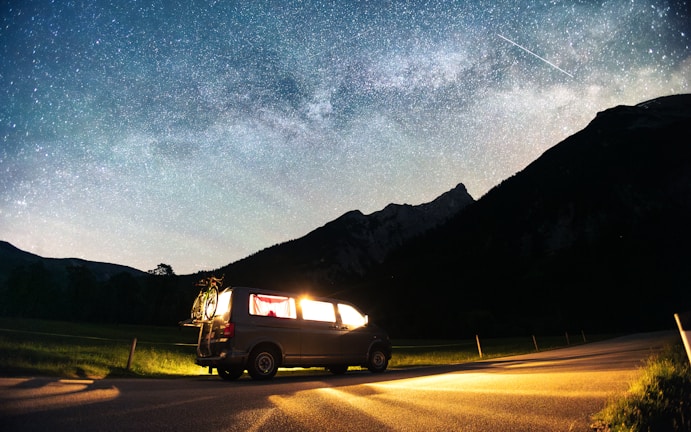 A camper van is parked on a roadside under a clear night sky filled with stars and a visible Milky Way. The van's interior lights are on, casting a warm glow, and a bicycle is attached to the back. Mountains create a silhouetted backdrop, adding to the serene and adventurous setting.