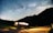 A camper van is parked on a roadside under a clear night sky filled with stars and a visible Milky Way. The van's interior lights are on, casting a warm glow, and a bicycle is attached to the back. Mountains create a silhouetted backdrop, adding to the serene and adventurous setting.
