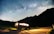 A camper van is parked on a roadside under a clear night sky filled with stars and a visible Milky Way. The van's interior lights are on, casting a warm glow, and a bicycle is attached to the back. Mountains create a silhouetted backdrop, adding to the serene and adventurous setting.