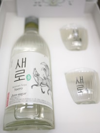 A bottle of Korean Soju labeled 'Saero' with an illustration of a fox on the label is placed inside a white box. Beside the bottle, there are two small, clear glasses, each featuring similar markings to the bottle. The bottle's label includes text indicating 'Zero sugar' and describes the product as distilled in Korea.