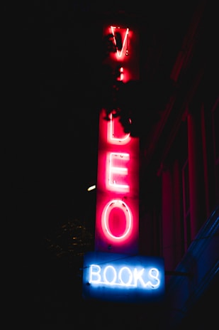 A neon sign glowing in the dark with the words 'VIDEO' and 'BOOKS'. The letters 'VIDEO' are vertically aligned in red, while 'BOOKS' is horizontally aligned in blue. The sign appears to be against a building facade.