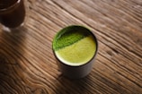 A cup filled with frothy green tea sits on a textured wooden surface. The green tea has a vibrant appearance with a fine, powdery layer on top, possibly matcha.