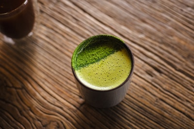 A cup filled with frothy green tea sits on a textured wooden surface. The green tea has a vibrant appearance with a fine, powdery layer on top, possibly matcha.