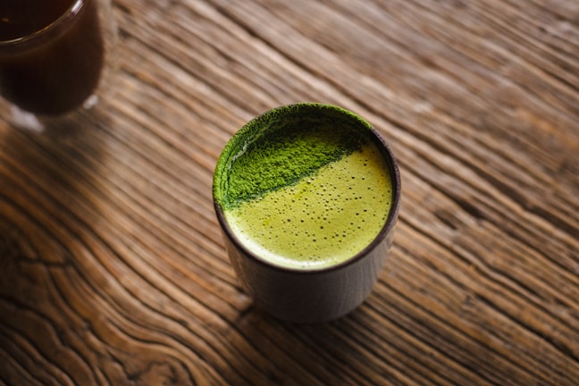 A cup filled with frothy green tea sits on a textured wooden surface. The green tea has a vibrant appearance with a fine, powdery layer on top, possibly matcha.