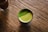 A cup filled with frothy green tea sits on a textured wooden surface. The green tea has a vibrant appearance with a fine, powdery layer on top, possibly matcha.