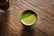 A cup filled with frothy green tea sits on a textured wooden surface. The green tea has a vibrant appearance with a fine, powdery layer on top, possibly matcha.