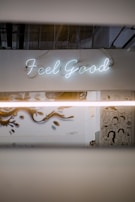 A neon sign displaying the words 'Feel Good' is mounted on a white wall. Below, there is a mural with artistic illustrations of people and stylized designs. The setting has an industrial vibe with visible ceiling pipes and muted lighting.