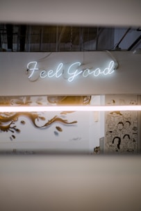 A neon sign displaying the words 'Feel Good' is mounted on a white wall. Below, there is a mural with artistic illustrations of people and stylized designs. The setting has an industrial vibe with visible ceiling pipes and muted lighting.