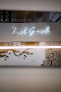A neon sign displaying the words 'Feel Good' is mounted on a white wall. Below, there is a mural with artistic illustrations of people and stylized designs. The setting has an industrial vibe with visible ceiling pipes and muted lighting.