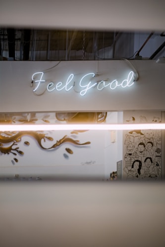 A neon sign displaying the words 'Feel Good' is mounted on a white wall. Below, there is a mural with artistic illustrations of people and stylized designs. The setting has an industrial vibe with visible ceiling pipes and muted lighting.
